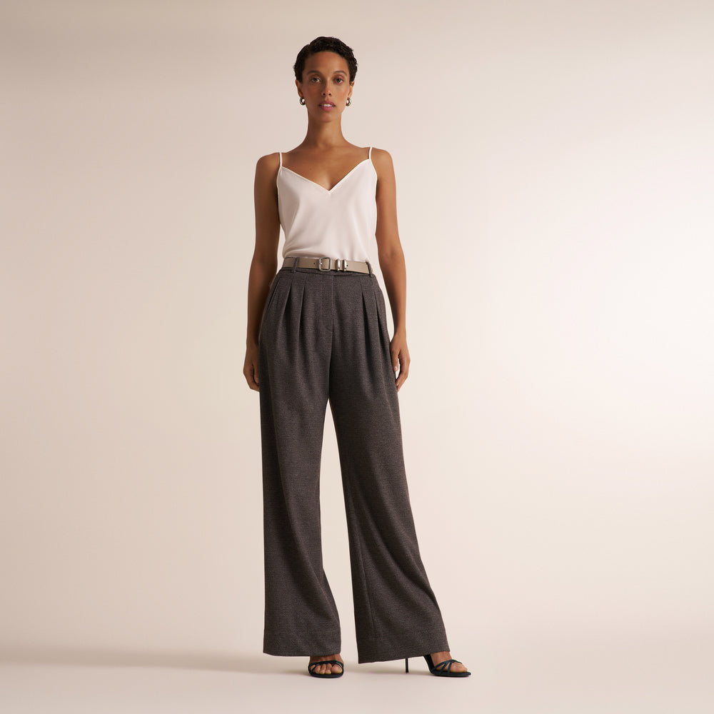 A model stands against a plain beige studio background wearing a white camisole top, dark charcoal gray wide-leg pleated trousers with a metallic silver belt, and black high-heeled sandals.