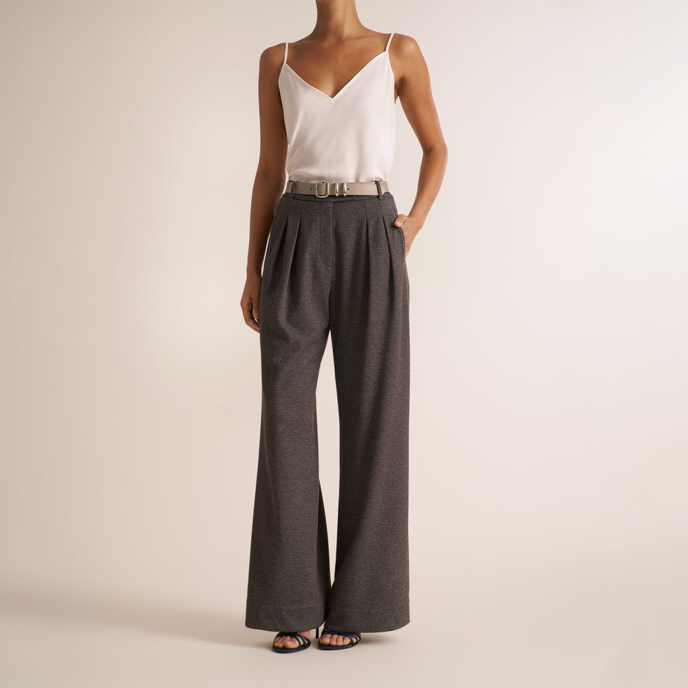 A model wearing dark gray wide-leg pleated trousers with a matching belt, paired with a white camisole top and black sandals, standing against a plain beige studio background.