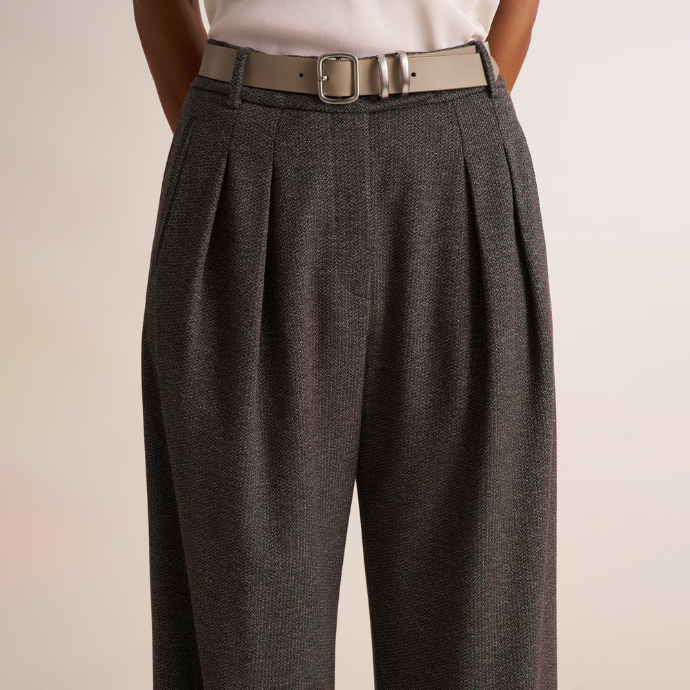 A close-up of a person wearing high-waisted, wide-leg charcoal gray pleated trousers with a beige leather belt and a white top, set against a plain light beige background.