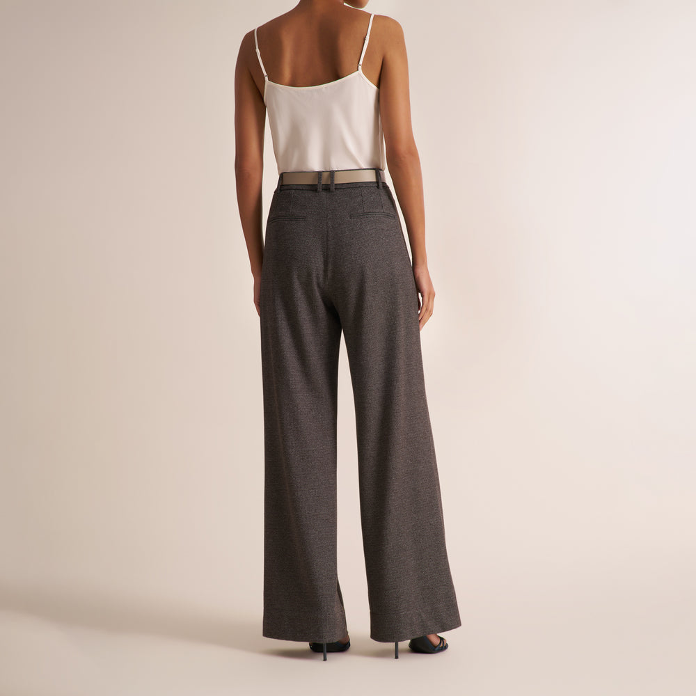 A model shown from the back wearing high-waisted, wide-leg charcoal gray trousers, a white spaghetti strap top, and a thin metallic belt against a warm beige studio background.