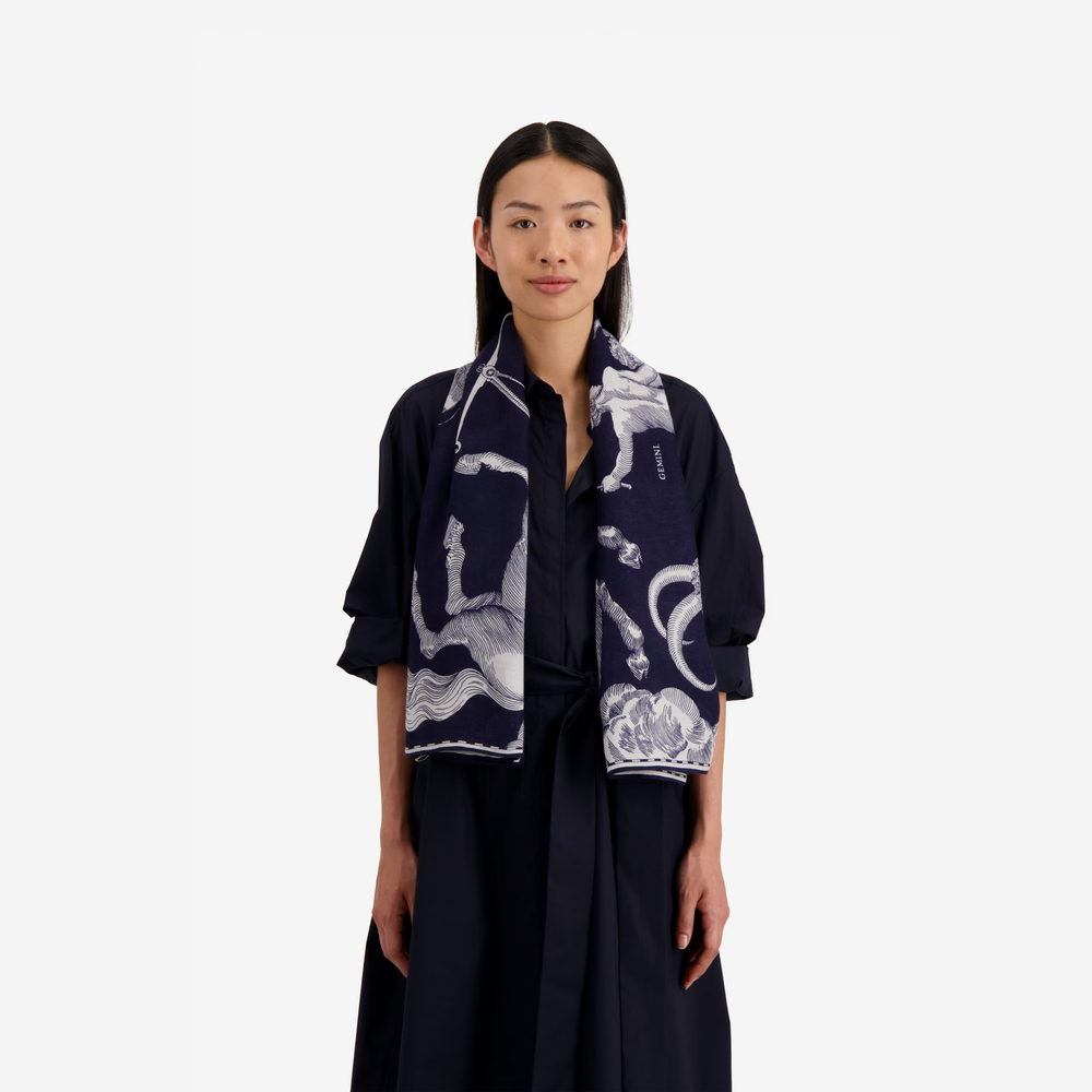 A model wearing a navy blue silk scarf with a white celestial print, styled over a dark navy dress against a solid light gray background.