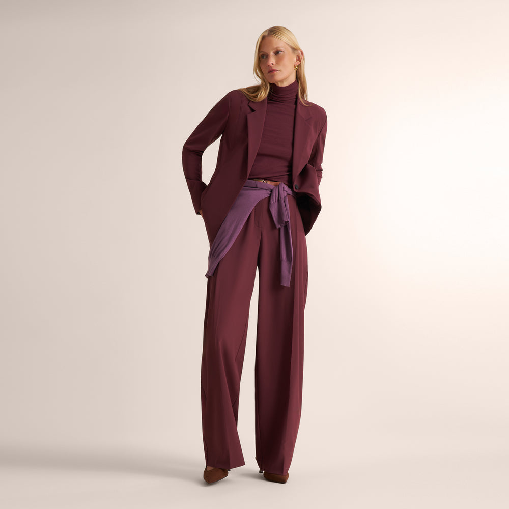 A woman models a coordinated outfit consisting of a deep burgundy blazer and matching turtleneck top, paired with wide-leg trousers in a matching burgundy fabric and a purple draped sash at the waist.