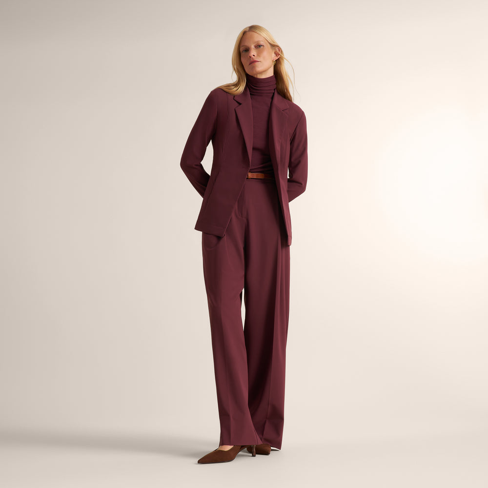 A woman modeling a tailored burgundy blazer and matching trousers with a matching turtleneck, cinched at the waist with a brown belt, against a plain light beige studio background.
