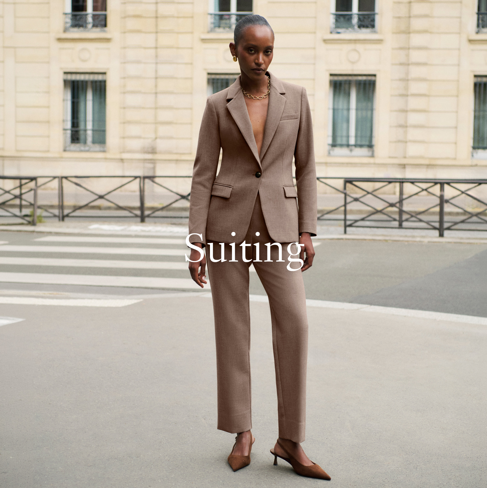 A model stands on a city street wearing a tailored taupe-brown suit with a single-breasted blazer and matching trousers, paired with brown pointed-toe heels. The background shows a classic beige stone building with windows and a crosswalk.

"Suiting" is overlaid