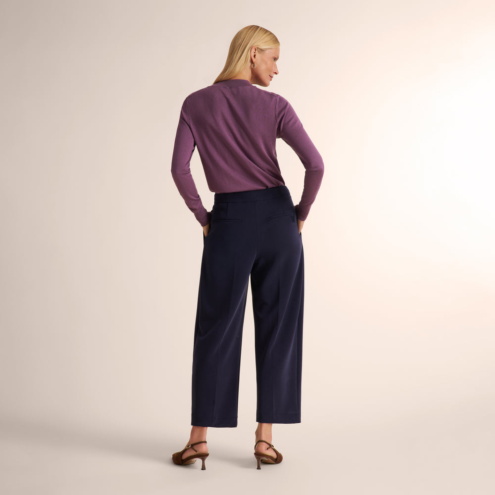 A woman stands with her back to the camera wearing a purple long-sleeve top and navy wide-leg cropped trousers, paired with brown strappy heels, against a light beige studio background.