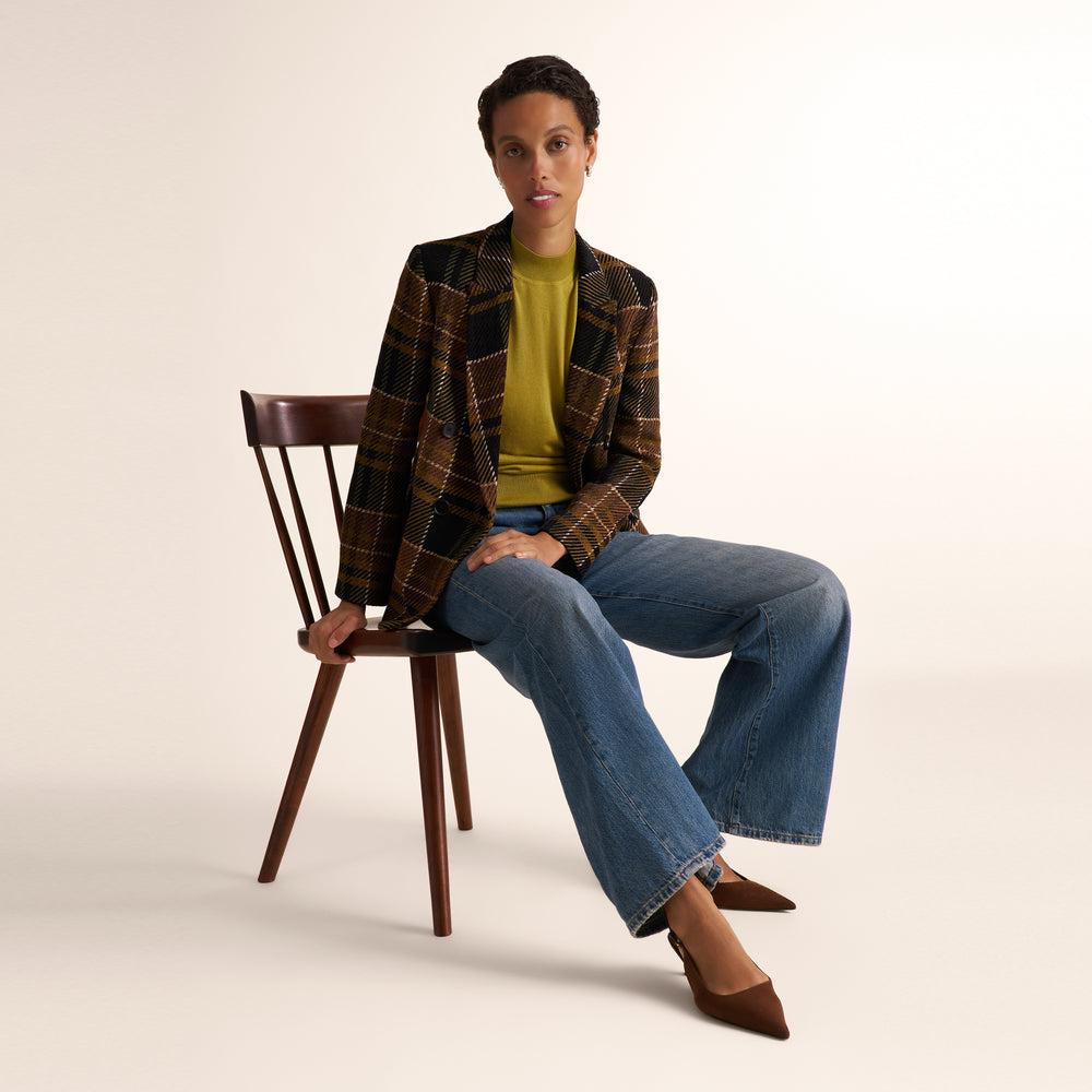 A model sits on a wooden chair wearing a brown and black plaid blazer over a mustard yellow top, blue wide-leg jeans, and brown pointed-toe heels against a plain off-white studio background.