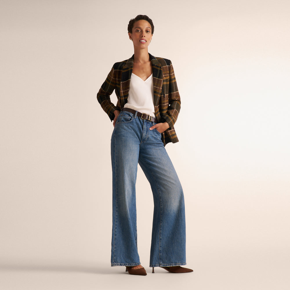 A model stands against a warm beige studio background wearing a brown and black plaid blazer over a white V-neck top, paired with wide-leg blue jeans and brown pointed-toe heels.