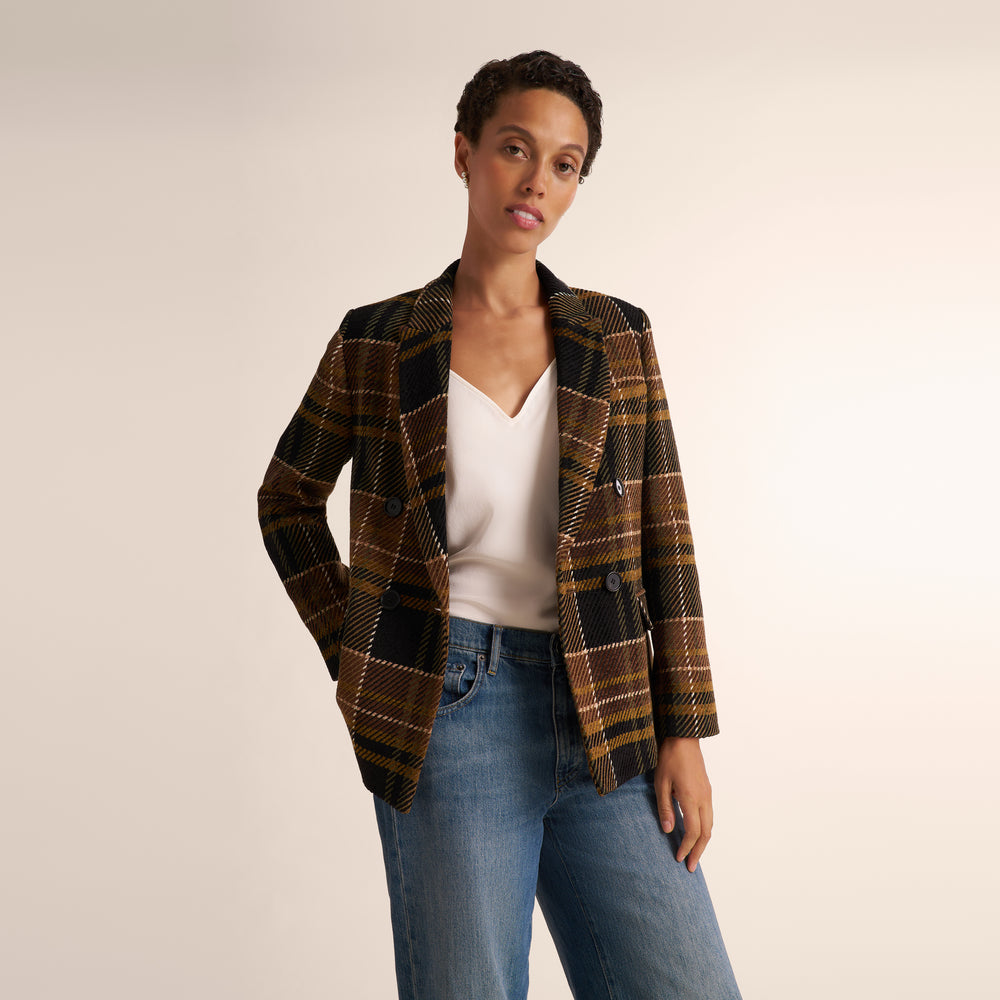 A model wearing a brown and black plaid blazer over a white top and blue jeans, standing against a plain beige studio background.