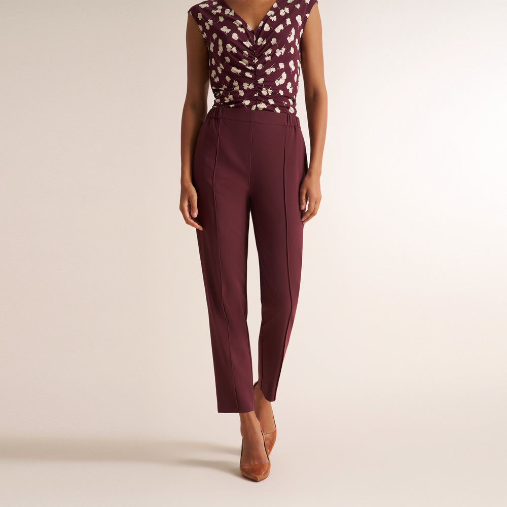 A model wearing a sleeveless burgundy top with a white floral pattern and matching high-waisted burgundy trousers, paired with brown pointed-toe heels, against a light beige studio background.