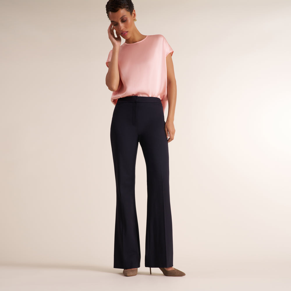 A model wearing a light pink short-sleeve top and dark navy flared trousers, standing against a plain beige studio background.