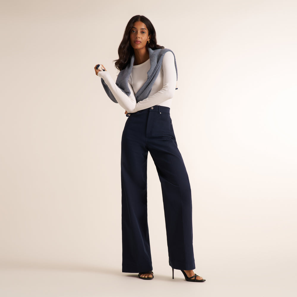 A model stands against a warm beige studio background wearing a pair of high-waisted, wide-leg navy trousers, a white long-sleeve top, a gray sleeveless vest, and black high-heeled sandals.