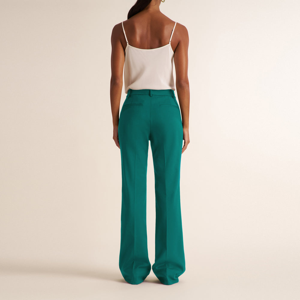 A woman shown from the back wearing high-waisted, wide-leg teal trousers and a white spaghetti-strap camisole against a warm beige studio background.