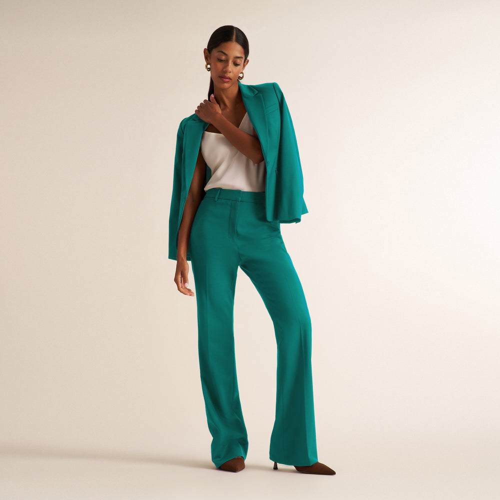 A model wearing a matching emerald green tailored set consisting of a draped open-front blazer and high-waisted wide-leg trousers, styled with a white top and brown pointed-toe heels against a warm beige studio background.