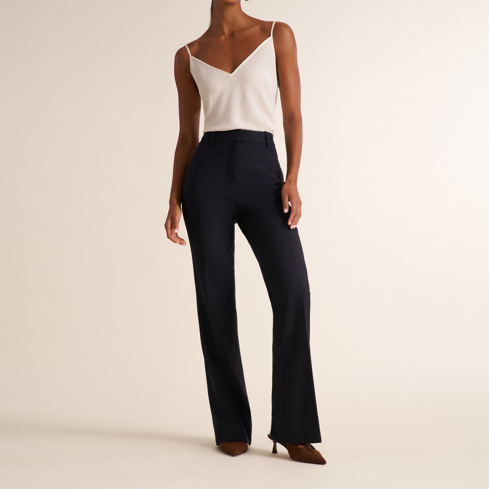 Woman wearing a black blazer, wide leg black suit pants, and a v-neck white tank top standing on a beige background