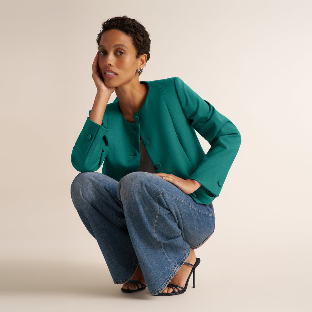 A model crouches in a studio setting wearing a teal green cropped blazer, wide-leg blue jeans, and black strappy heels against a solid light beige background.