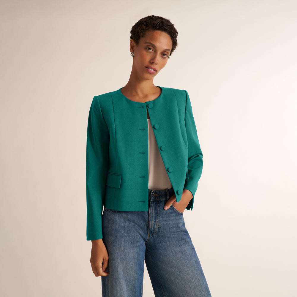 A woman modeling a teal green cropped jacket with a button front, worn over a light beige top and blue jeans, against a plain light beige studio background.