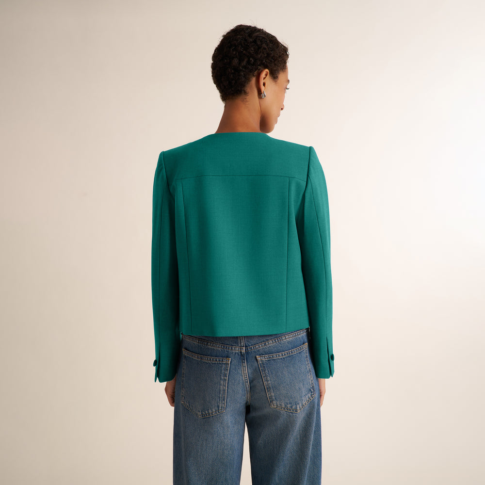A woman viewed from the back wearing a teal green long-sleeve cropped blazer and blue jeans, standing against a plain light beige studio background.