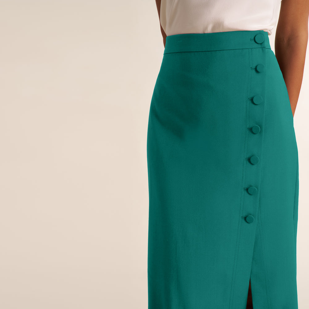 A close-up of a model wearing a high-waisted teal green skirt with a vertical row of large matching buttons down the front, paired with a white top against a warm beige background.