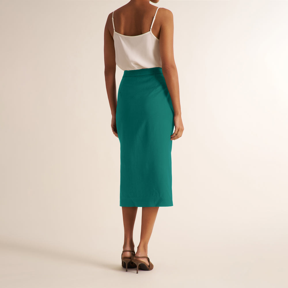 A woman shown from the back wearing a teal green midi skirt with a white spaghetti-strap top and brown strappy heels, set against a plain beige studio background.