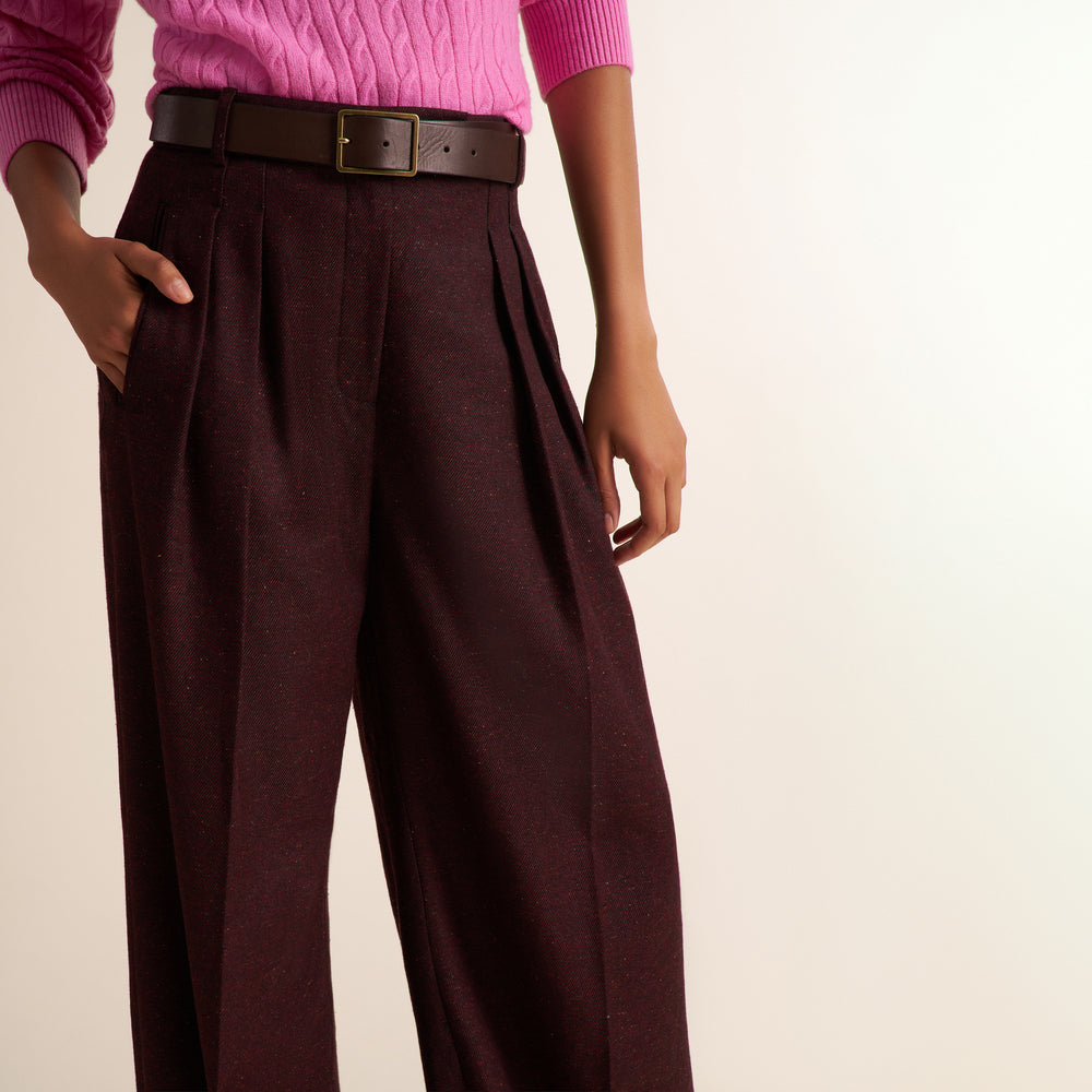 A person wearing high-waisted, wide-leg pleated trousers in a deep burgundy-brown color, styled with a dark brown leather belt and a pink sweater against a plain light background.