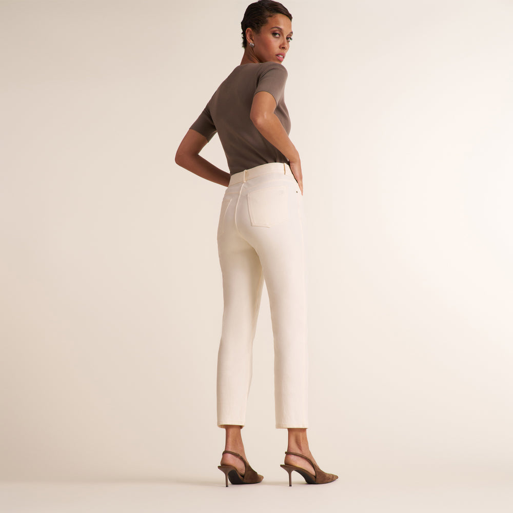 A woman stands in profile wearing off-white cropped pants, a taupe short-sleeve top, and brown heeled sandals against a plain warm beige studio background.