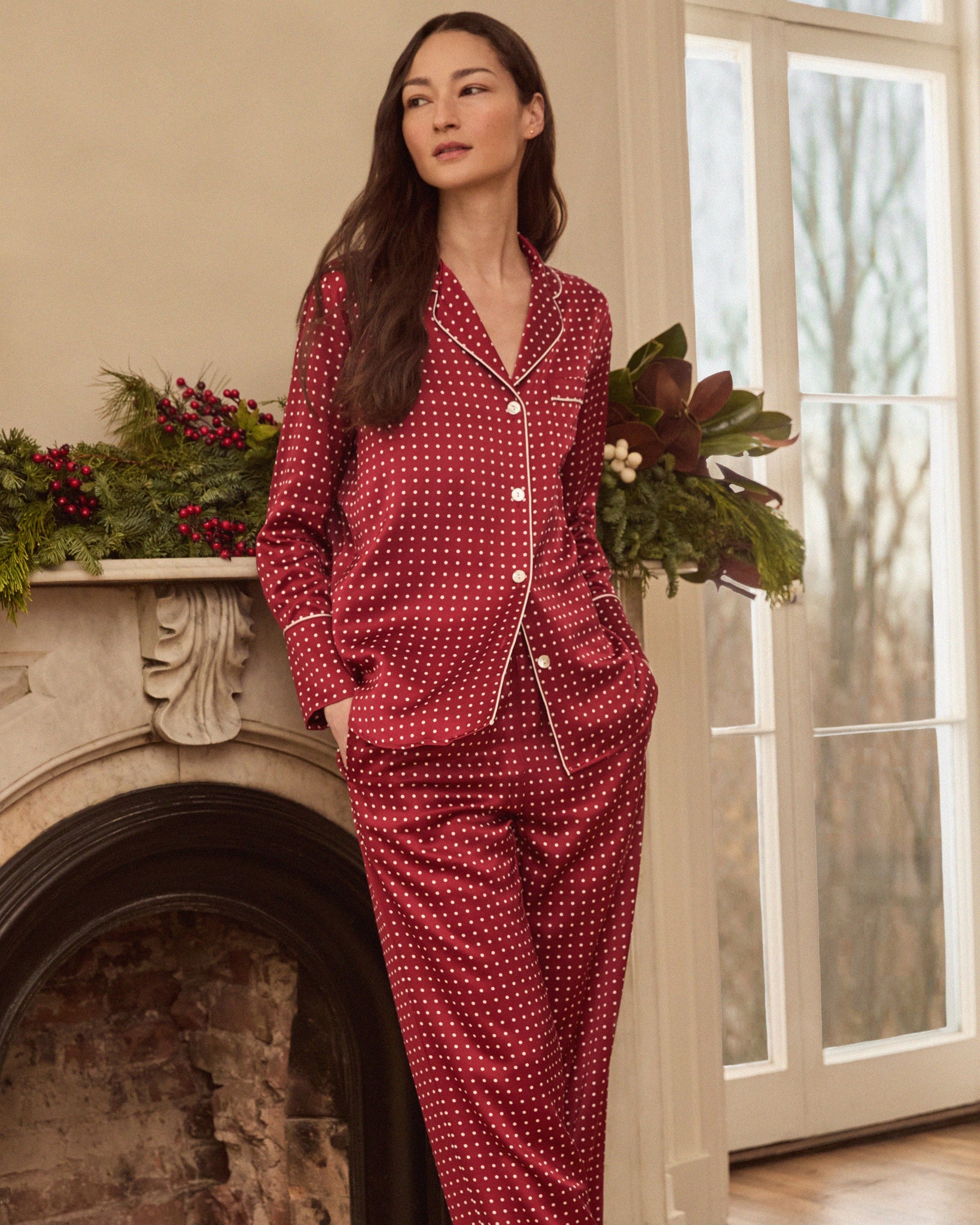 Woman wearing red polka dot pajamas standing by a fireplace with decorative plants.
