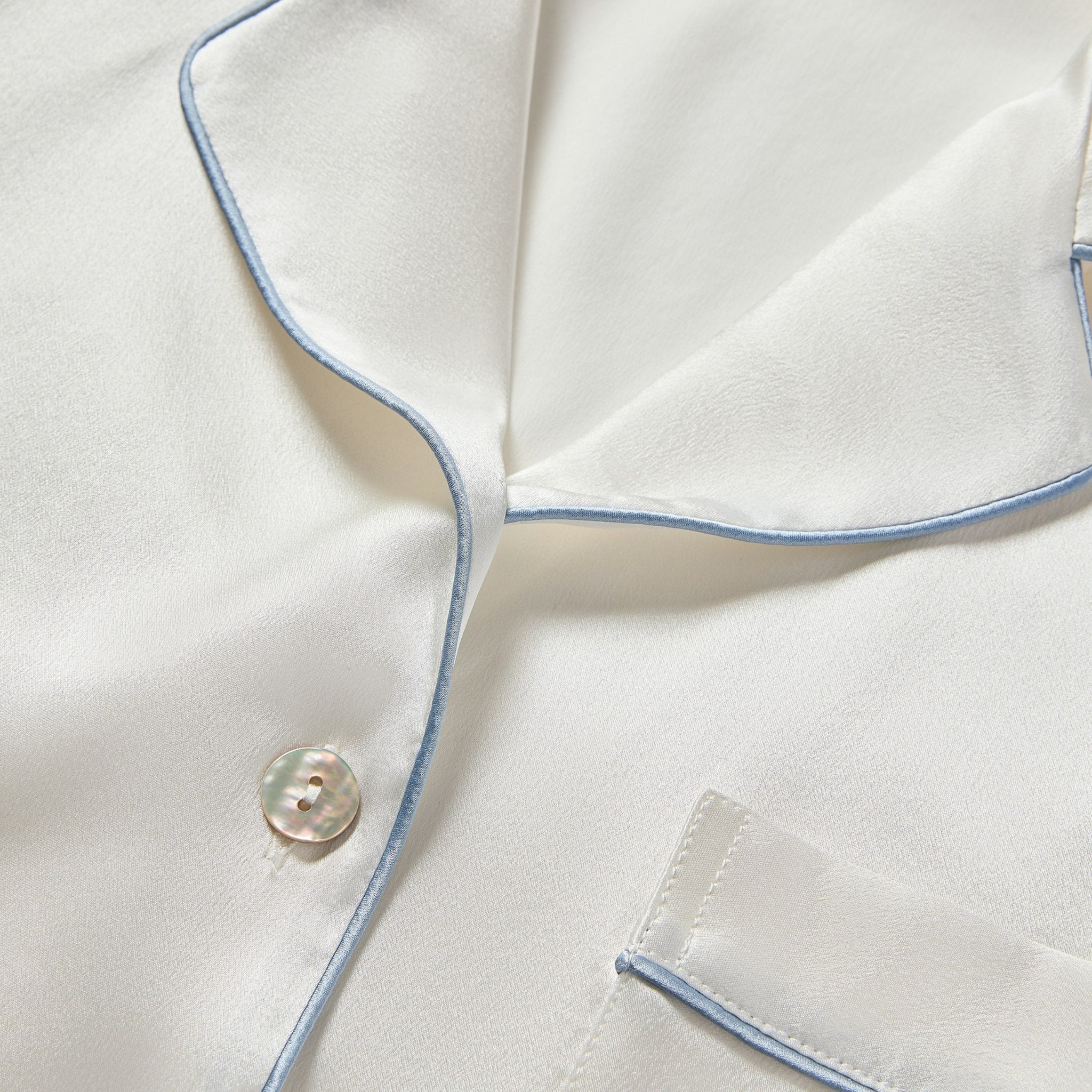 Close-up of a white shirt with blue piping and a button, focusing on the collar and chest pocket.