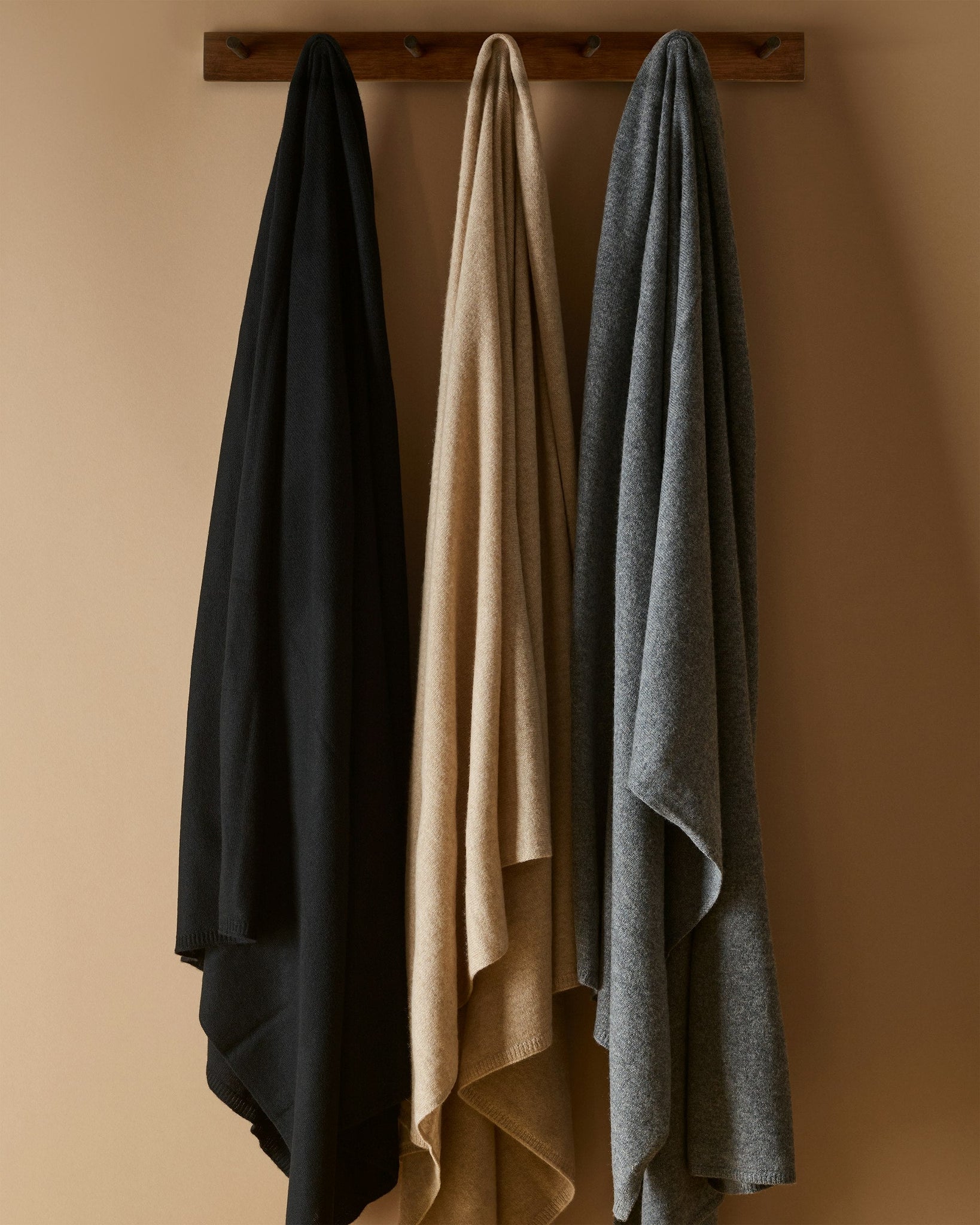 Three cloaks in black, beige, and gray hanging on a wooden rack against a beige wall.