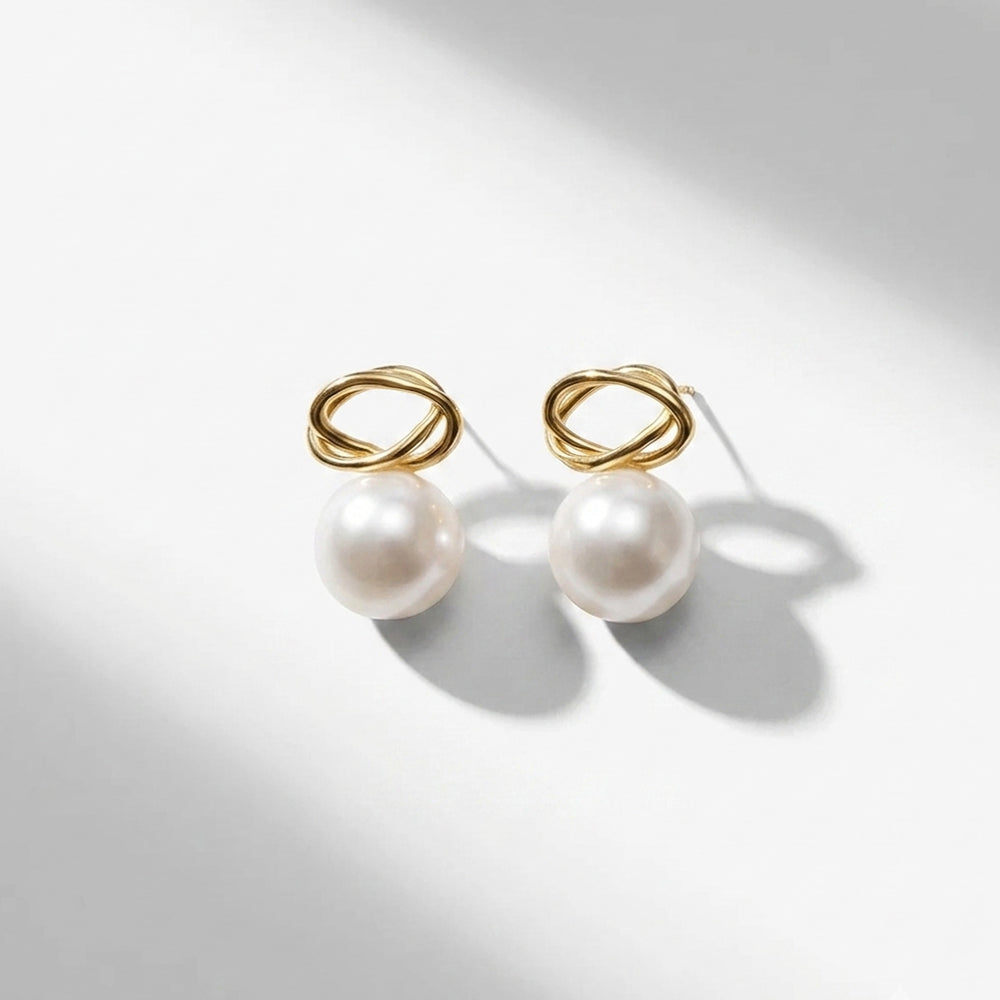 A pair of gold infinity loop stud earrings with a single white pearl hanging below each loop, photographed on a clean white surface with soft diagonal shadows.