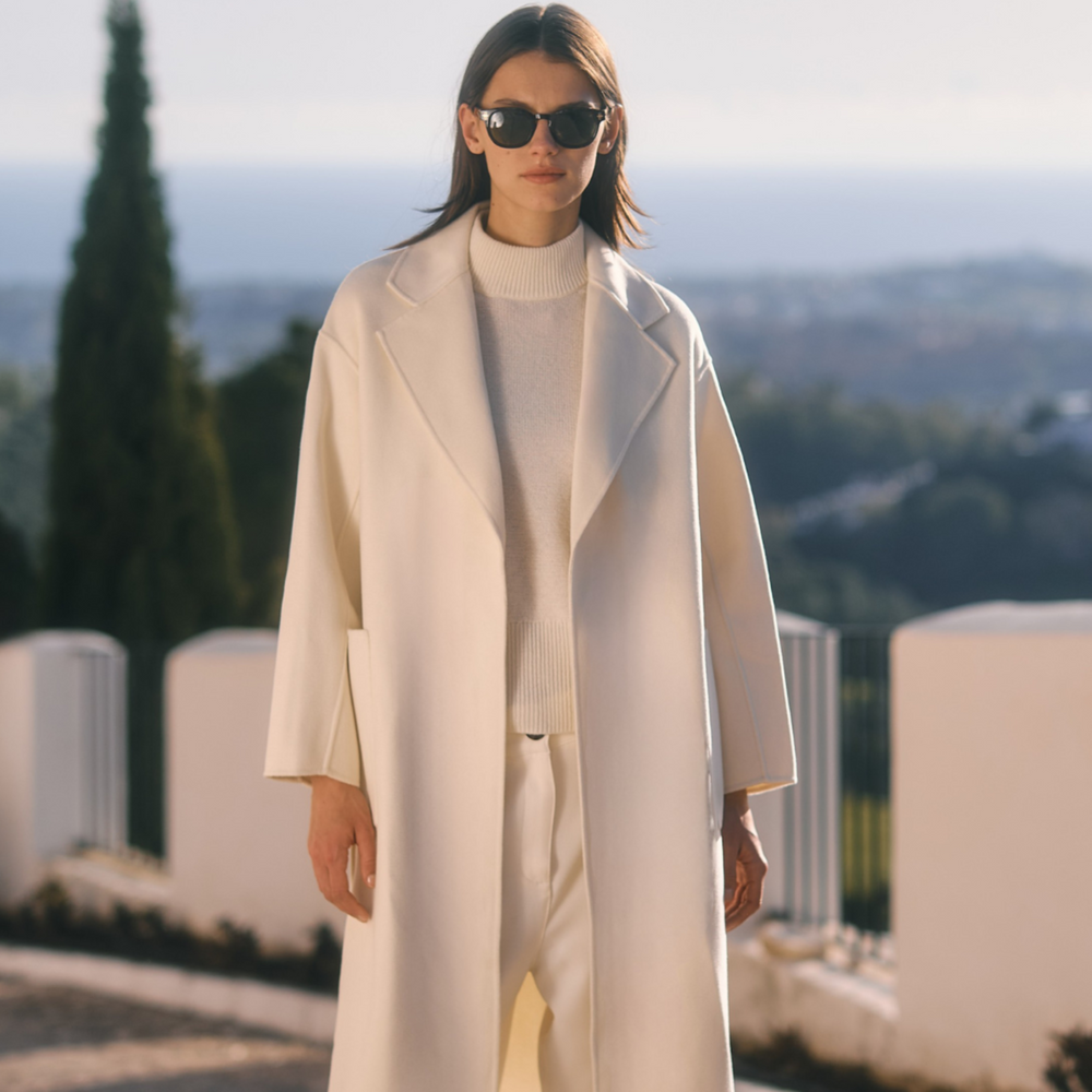 Person wearing a white coat and sunglasses in an outdoor setting with trees and buildings in the background