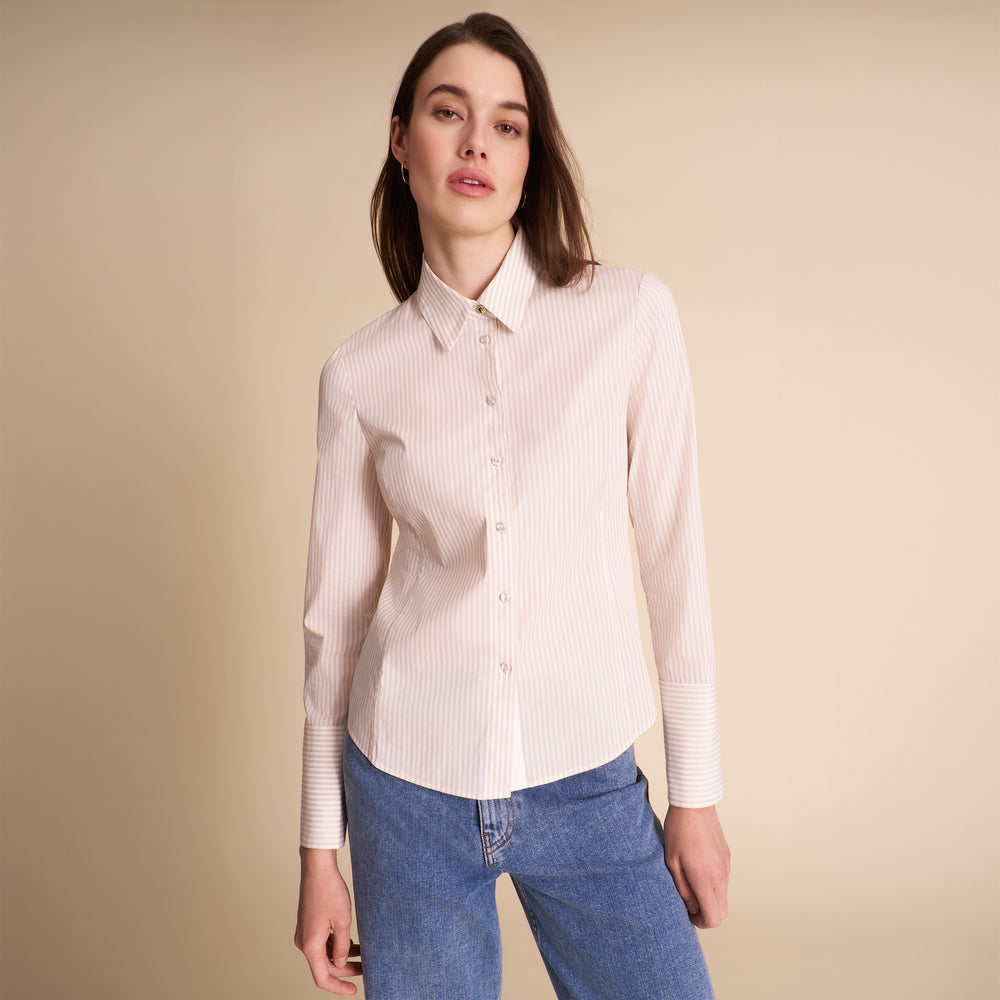 A woman modeling a light pink and white vertically striped button-down shirt with blue jeans, set against a solid beige studio background.