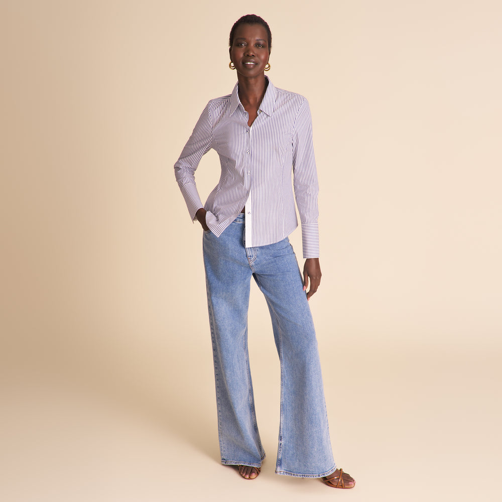 A woman modeling a light lavender and white vertically striped button-down shirt paired with light-wash blue flare jeans, standing against a solid beige studio background.