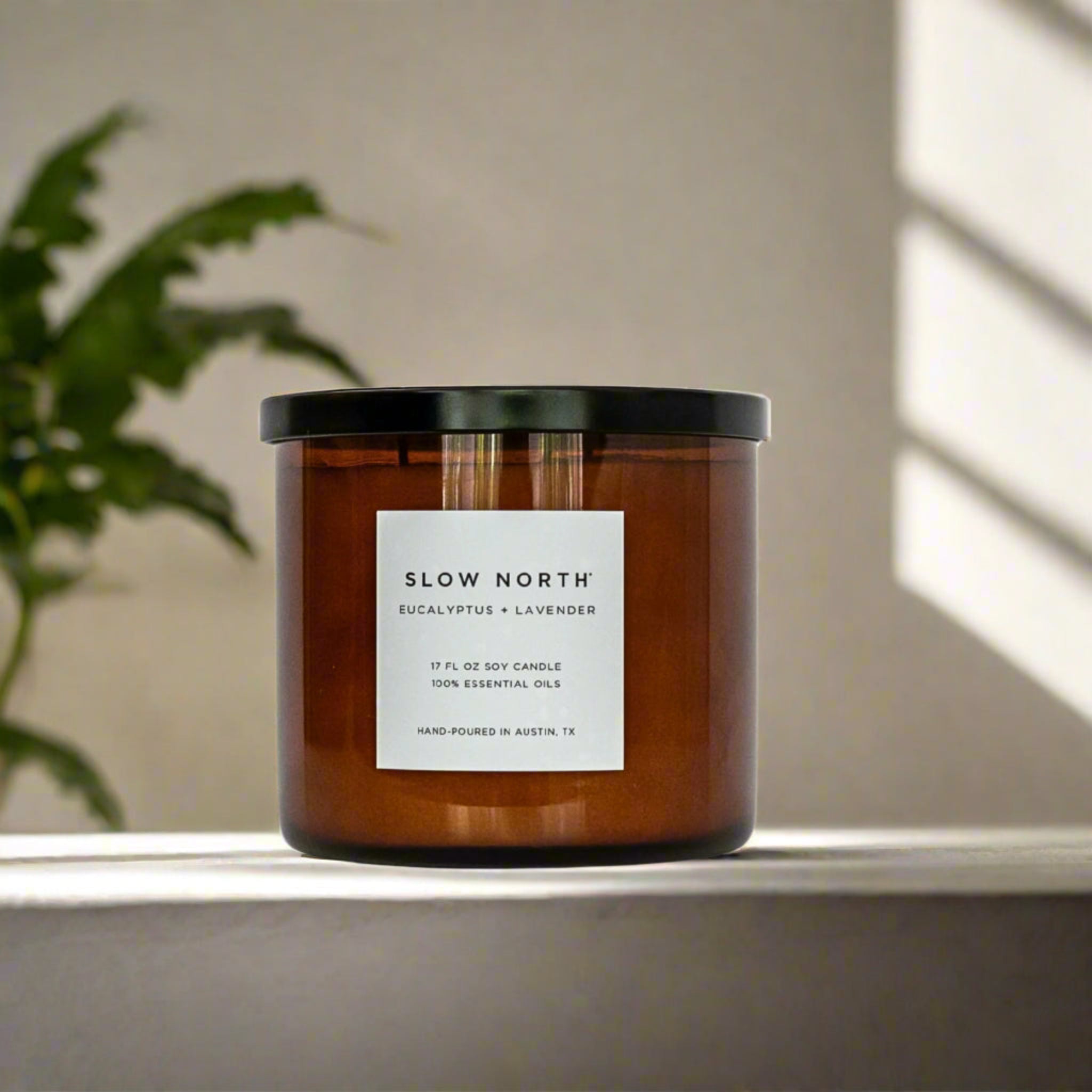 Candle with 'Slow North' label on a surface with a plant and light in the background