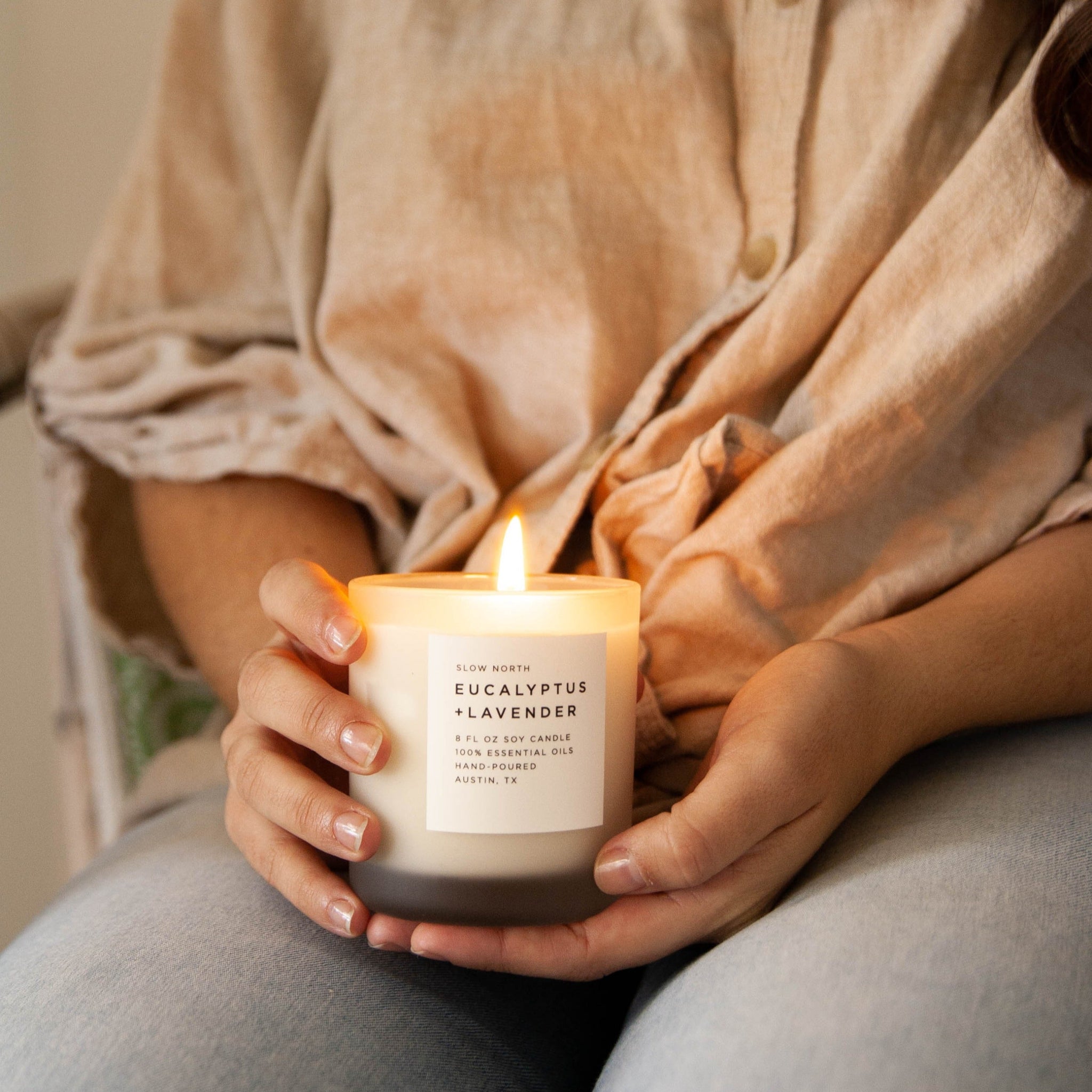Person holding a lit candle labeled 'Eucalyptus + Lavender' with a warm, cozy setting.