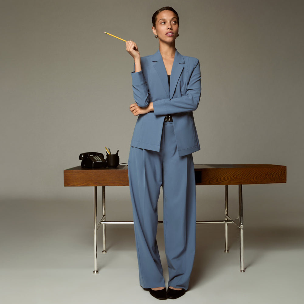 Woman in a blue suit holding a yellow pencil, standing next to a wooden desk with office items.