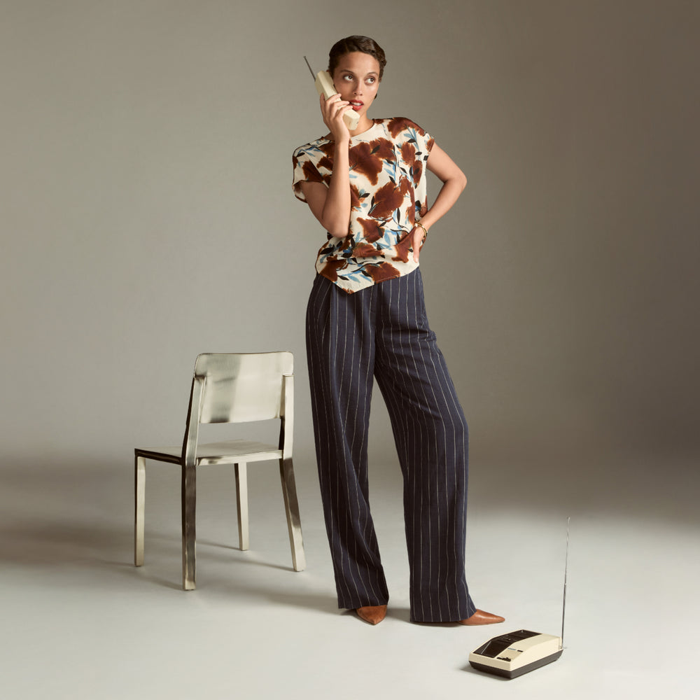 Woman in a patterned top and striped pants standing next to a chair and old-fashioned phone on a plain background