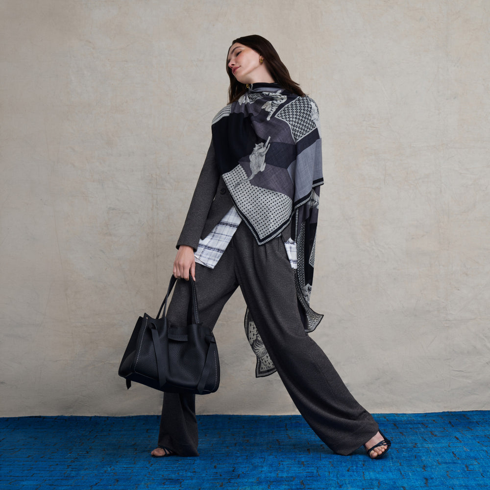 A woman models a stylish outfit featuring a black and gray patchwork scarf with geometric patterns, paired with a gray long-sleeve top and wide-leg charcoal trousers. She holds a black leather tote bag against a textured beige wall and a bright blue floor.