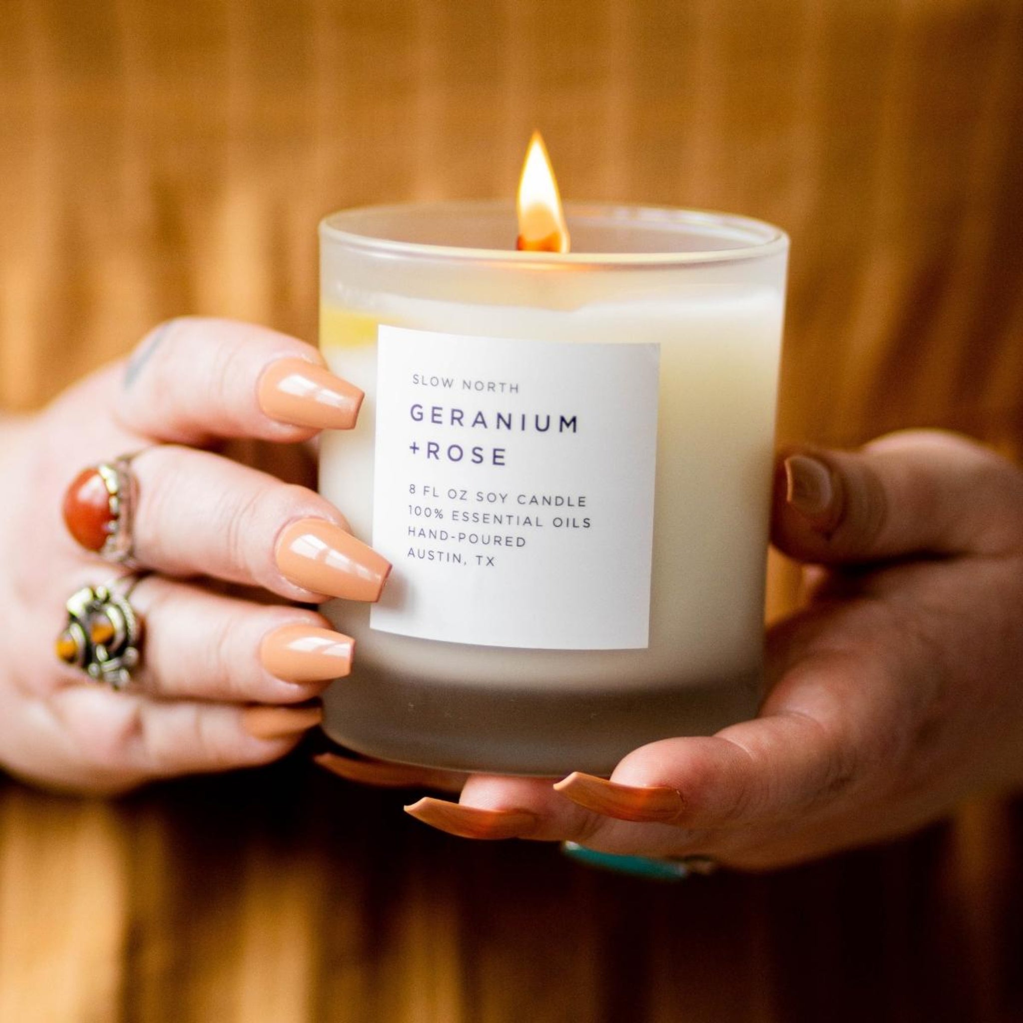 Candle labeled 'Geranium + Rose' held by hands with a blurred background