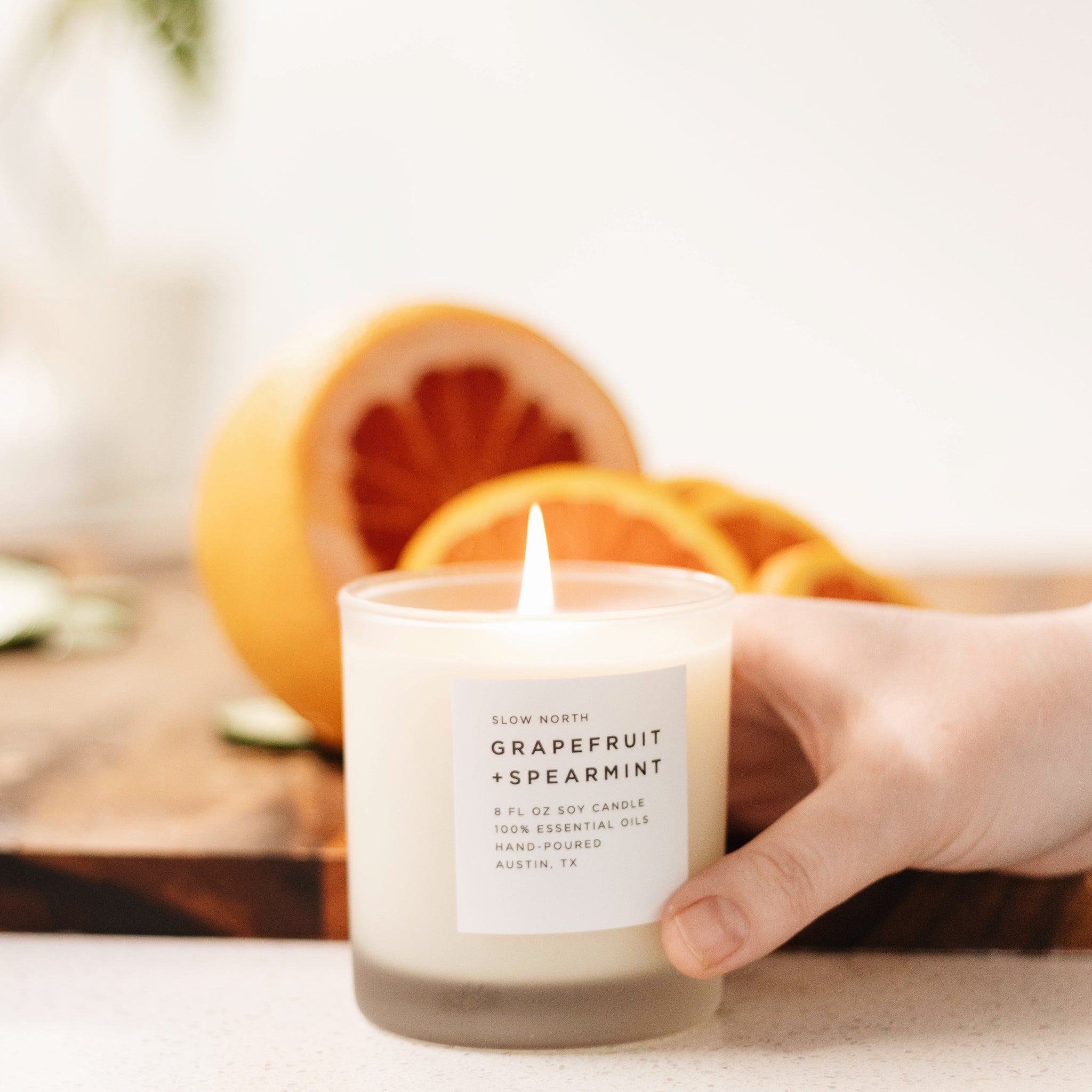 Candle labeled 'Grapefruit + Spearmint' held by a hand with oranges in the background