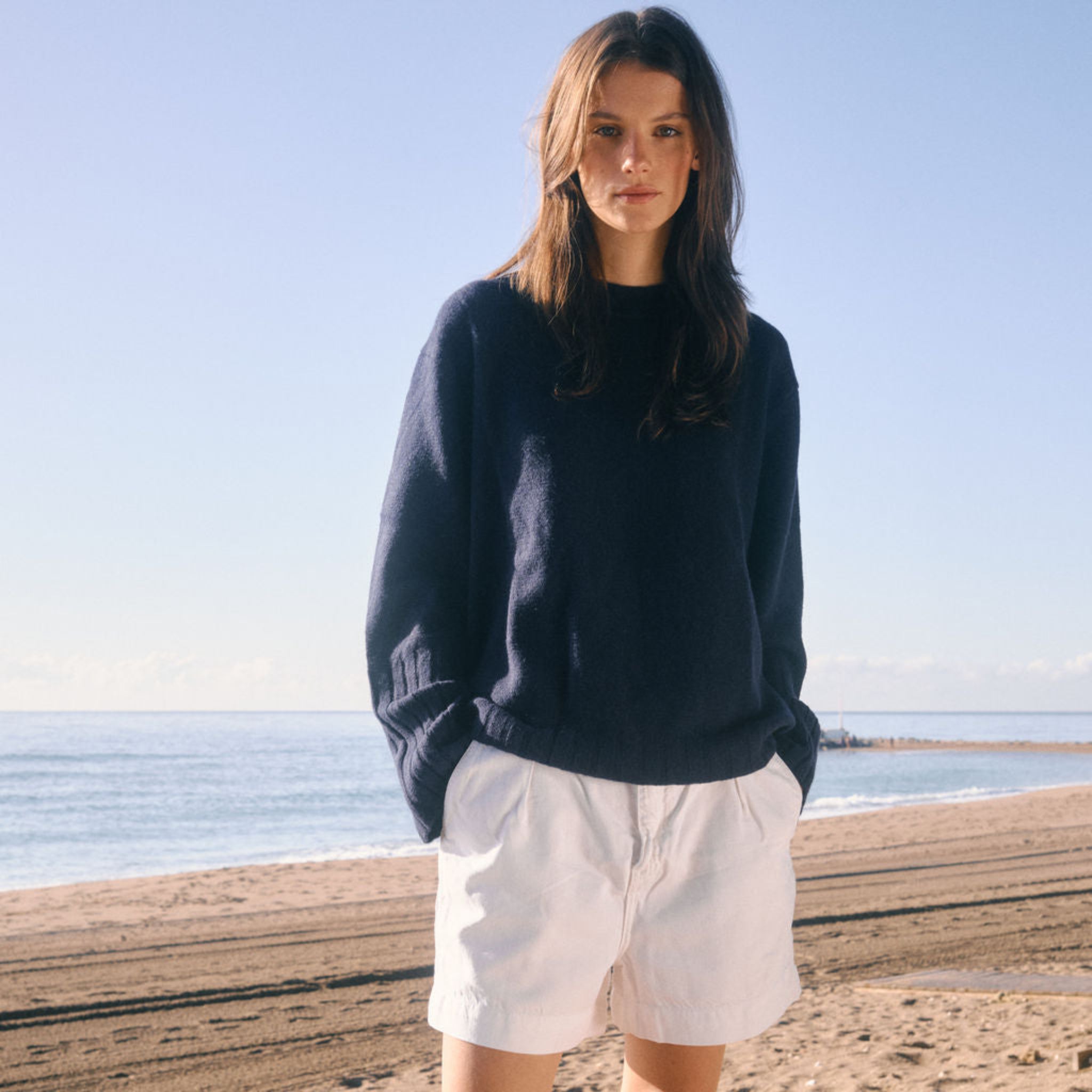 Woman standing on a beach wearing a navy sweater and white shorts.