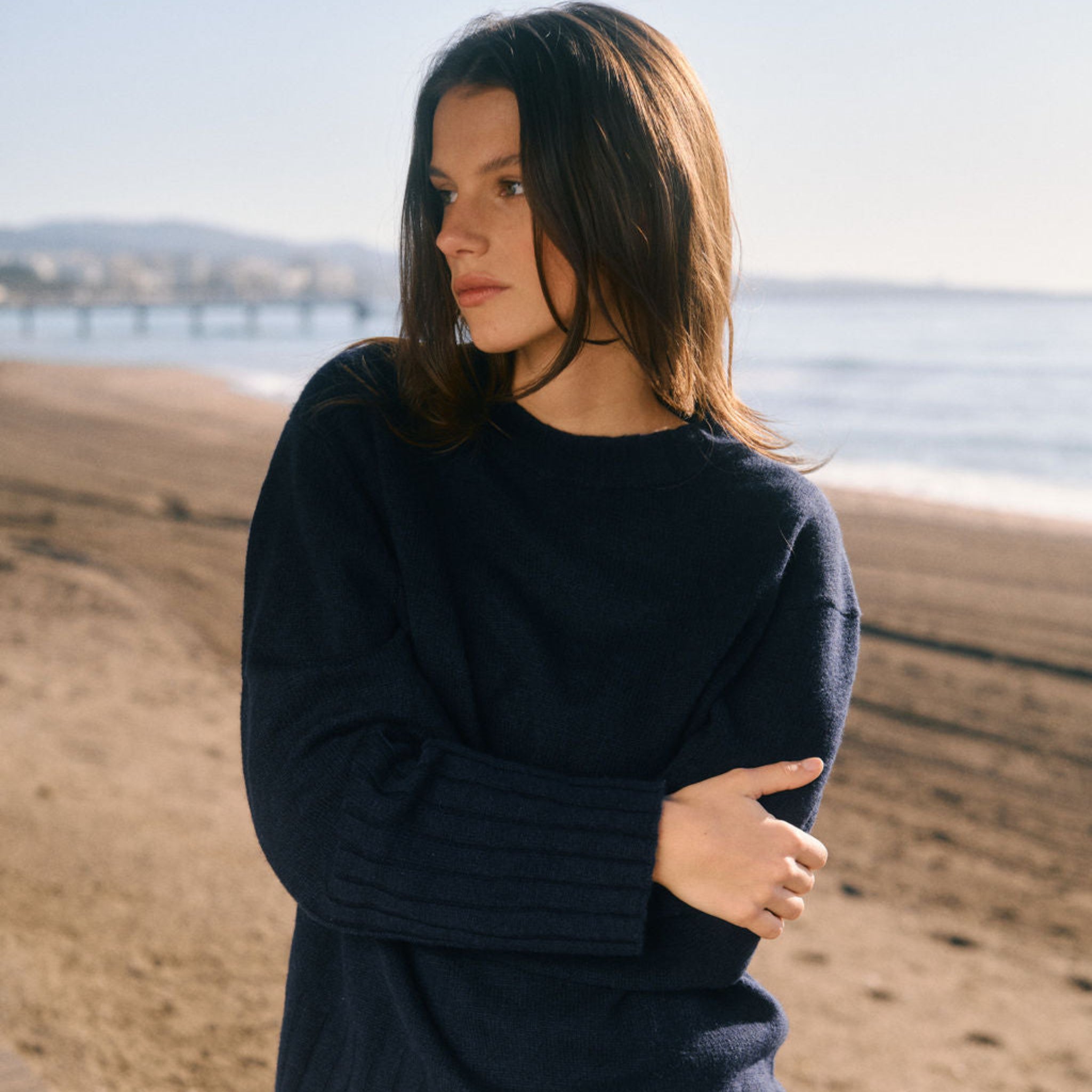 Woman wearing a navy sweater and white shorts standing on a sandy beach. 