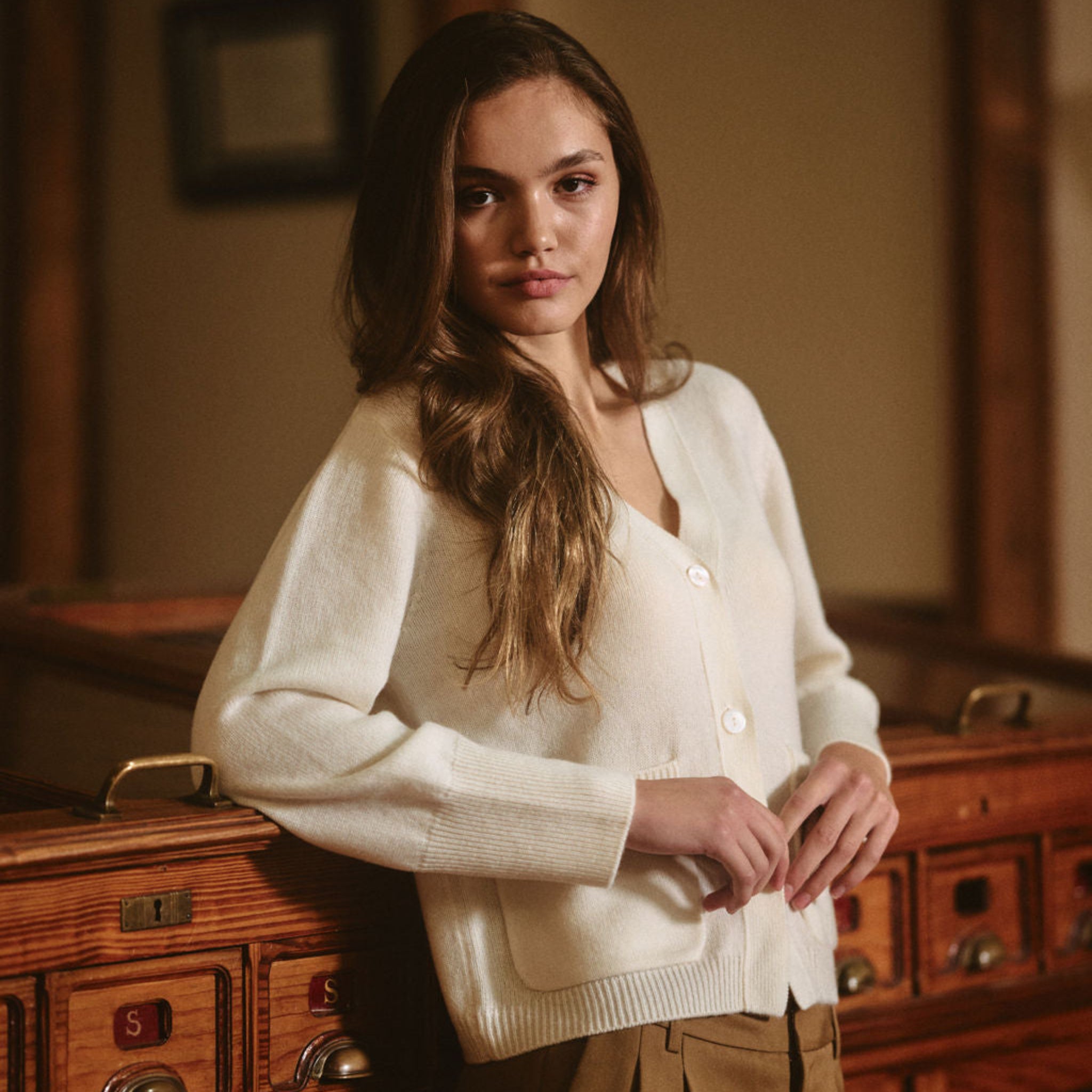 Woman wearing a white cardigan over a beige top in a room with wooden furniture. 