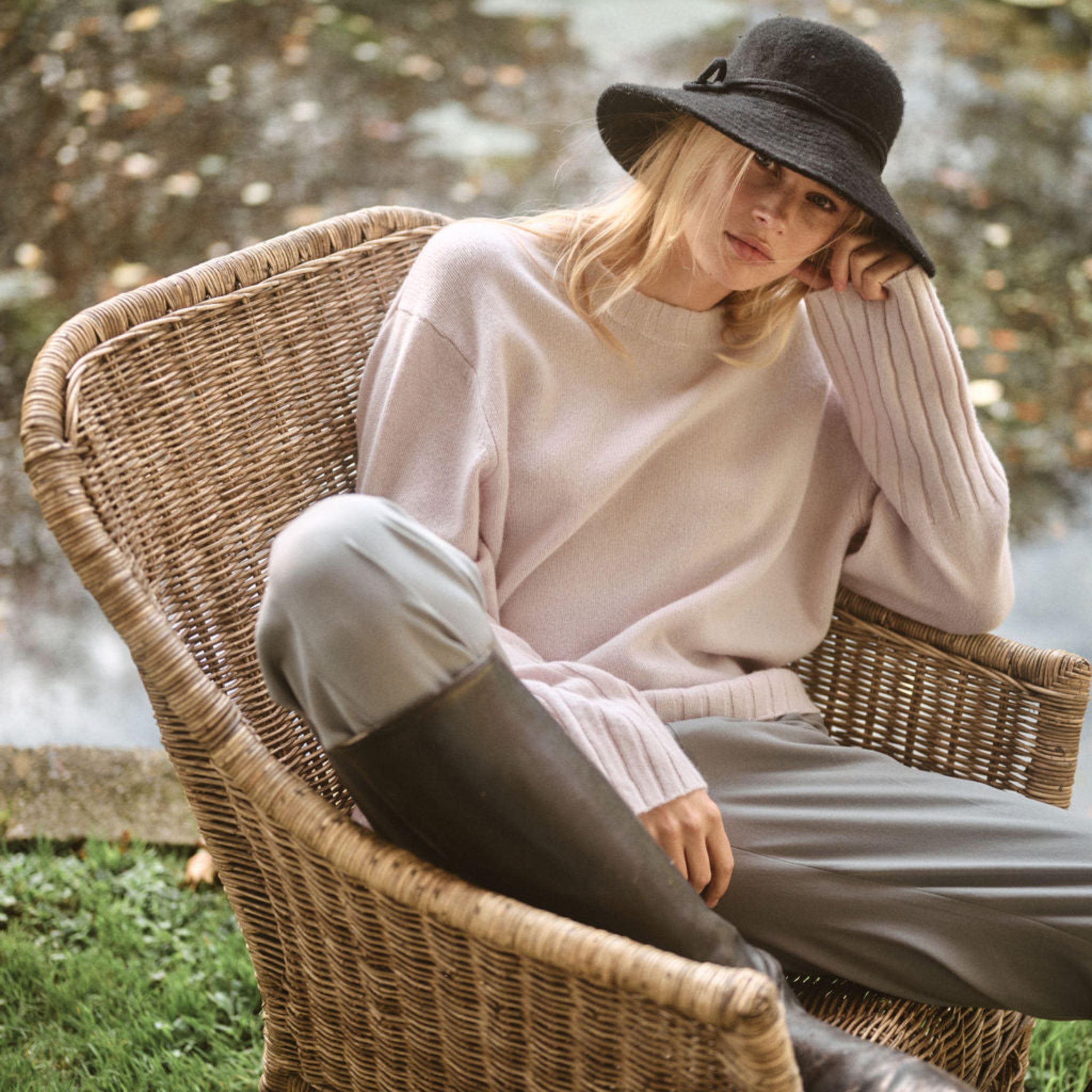 Woman sitting on a wicker chair by a body of water wearing a black hat and light-colored pants. 