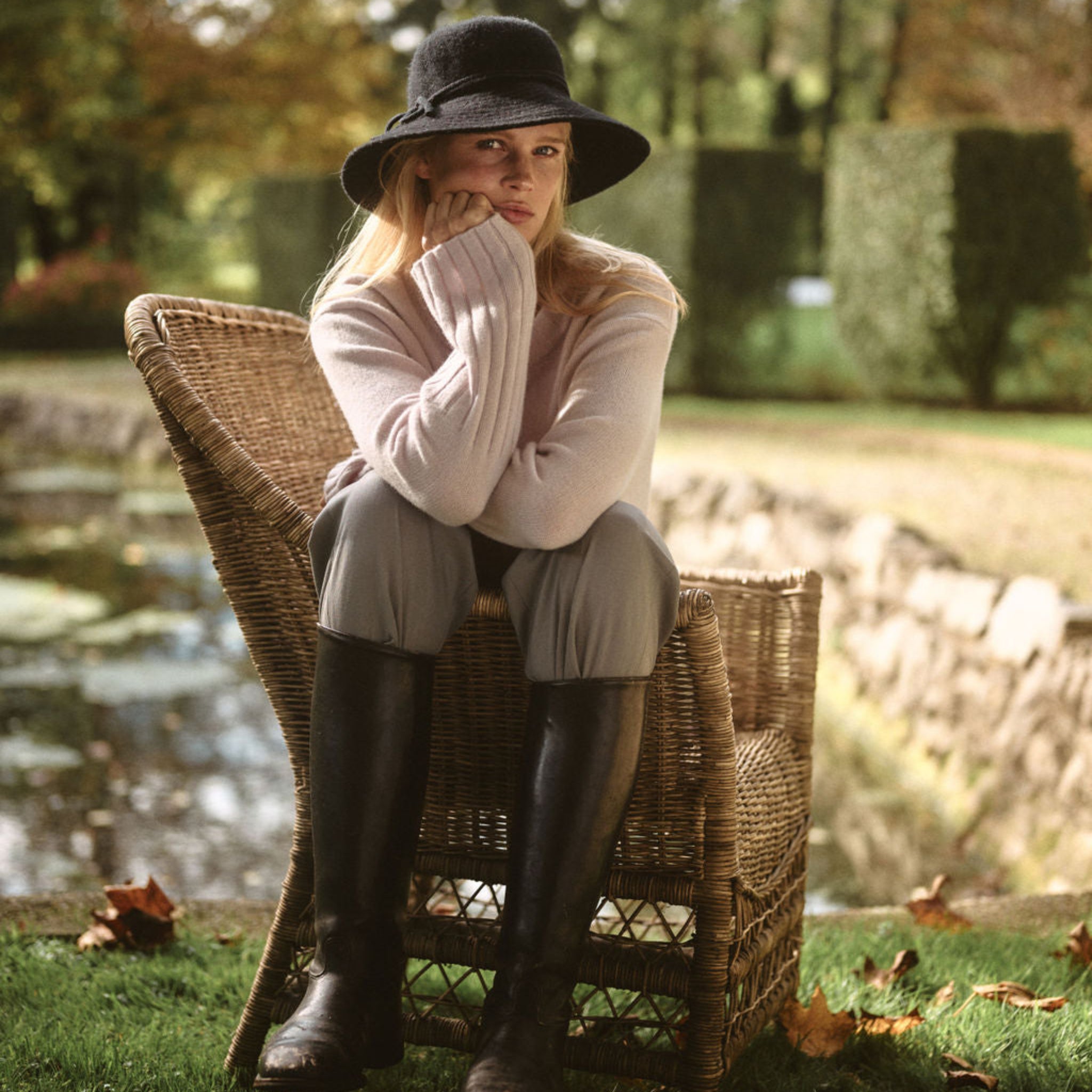 Woman sitting on a wicker chair by a pond wearing a hat, sweater, and boots.