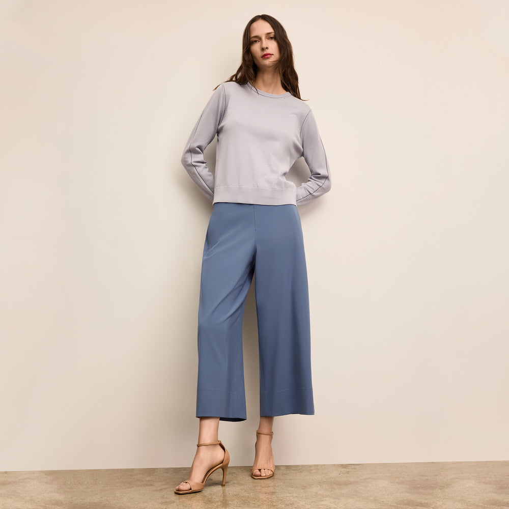 Woman wearing a light gray sweater and blue pants against a beige wall
