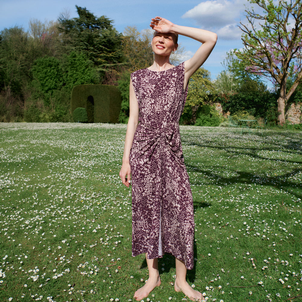 Woman in a purple floral dress standing in a garden with green grass and white flowers.