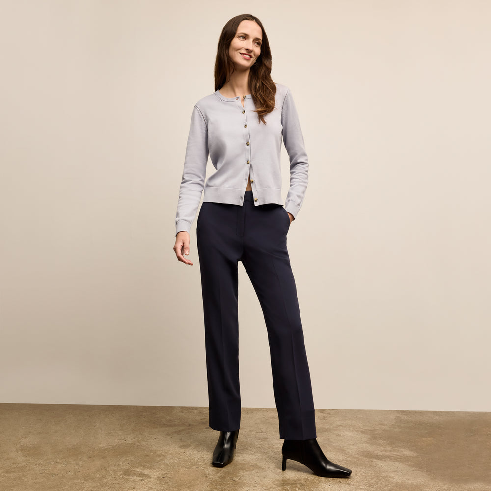 Woman wearing a light gray cardigan, black pants, and black shoes on a beige background