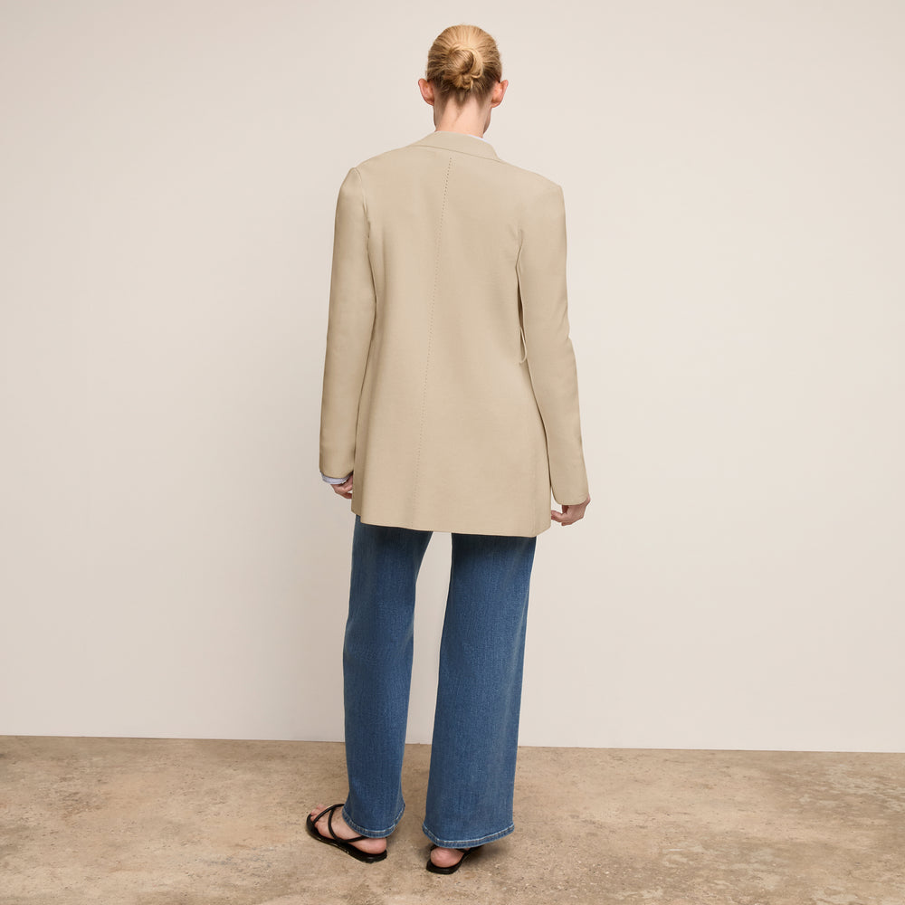 Person wearing a beige blazer and blue jeans standing against a plain background
