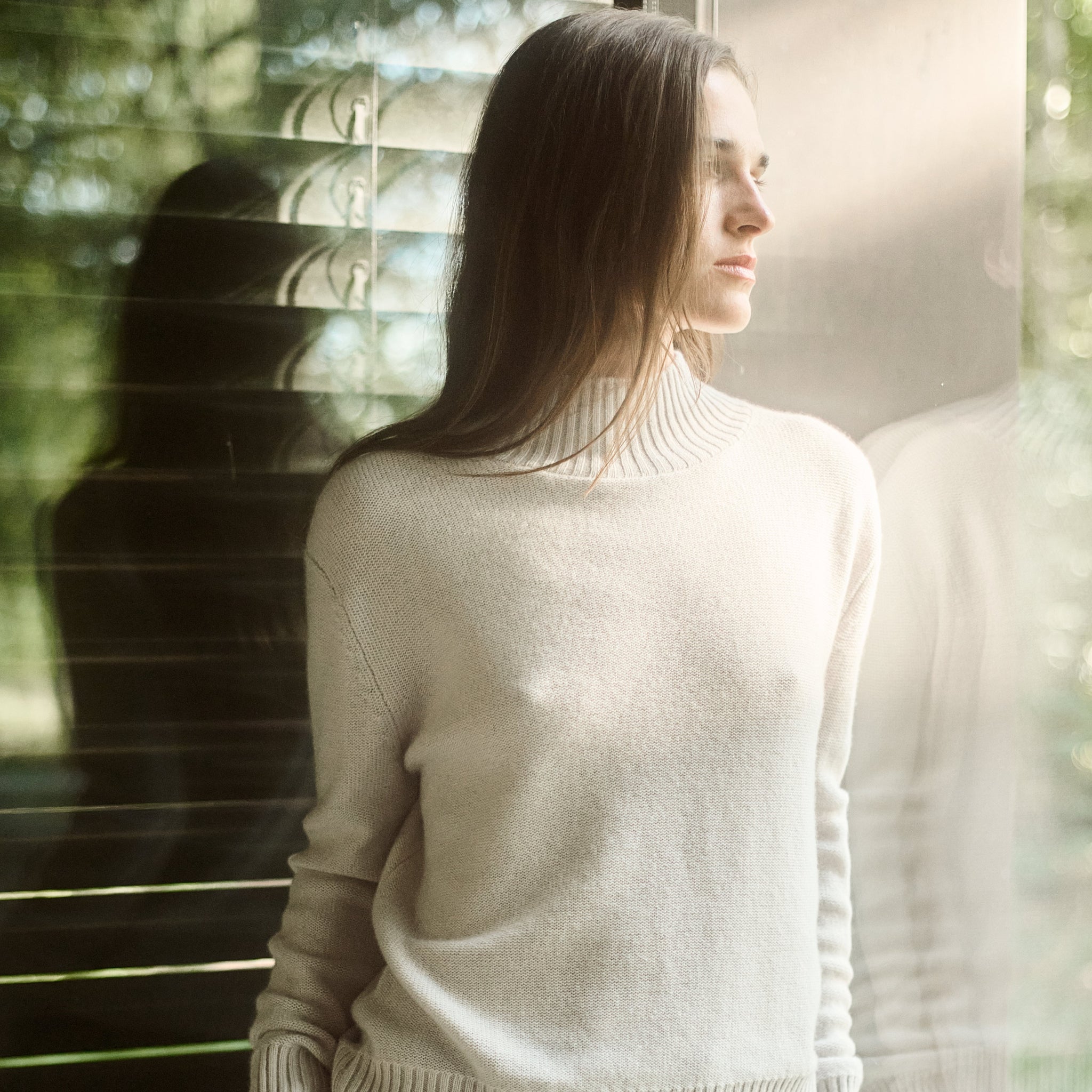 Woman wearing a beige sweater and dark pants standing by a window with a blurred outdoor background