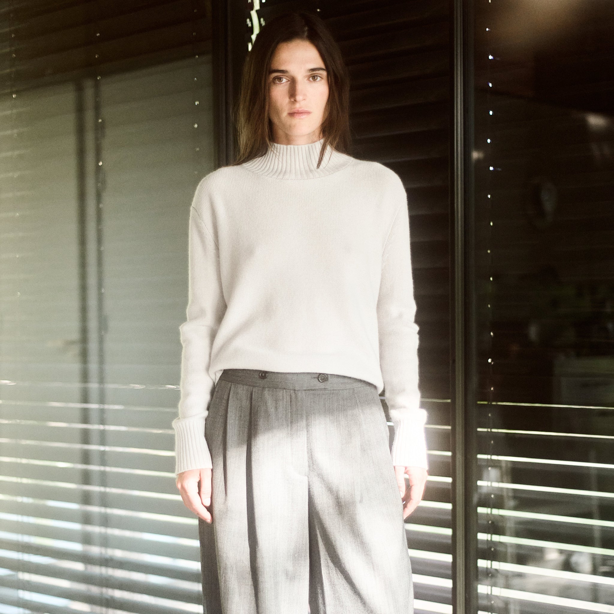 Person wearing a light-colored sweater and gray pants standing in front of a window with blinds.